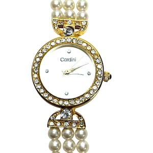 Peut inclure: Montre-bracelet dorée avec un cadran blanc et la marque "Cardini". La lunette et les détails sont incrustés de cristaux transparents. Le bracelet est composé de trois rangées de fausses perles.