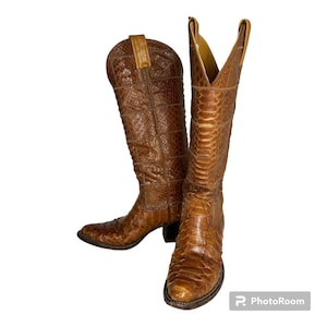 Könnte beinhalten: Ein Paar braune Leder-Cowboystiefel. Die Stiefel haben ein strukturiertes, gemustertes Design und eine spitze Zehenpartie. Die Stiefel sind kniehoch mit einem gestapelten Absatz. Das Leder hat eine warme, mittelbraune Farbe.