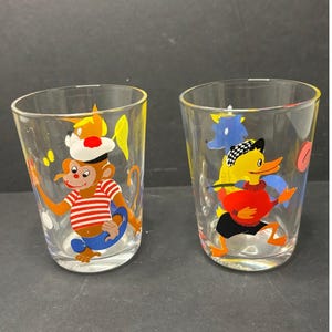May include: Two clear drinking glasses with cartoon animal designs. One glass features a monkey in a sailor outfit, and the other has a duck wearing a hat and sweater. The glasses have a yellow rim.