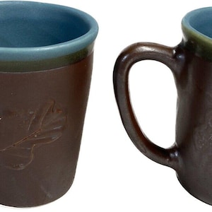 Puede incluir: Dos tazas de cerámica marrón con diseño de hojas. Las tazas tienen interior azul y borde verde. Las asas son curvas y las tazas ligeramente cónicas. Adecuadas para café, té u otras bebidas.