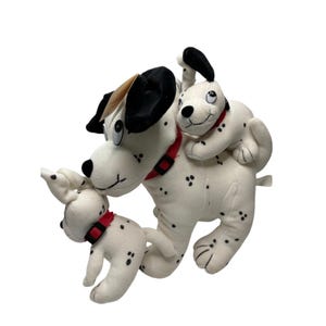 May include: A soft toy Dalmatian dog with two smaller puppies. The large dog has black spots, black ears, and a red collar. One puppy is on the dog's back, and the other is in front. The toys are white with black spots.