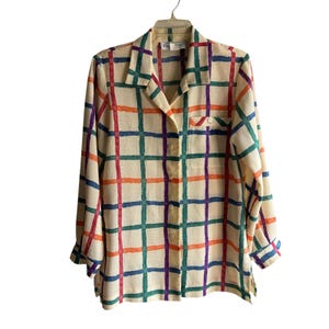 May include: A long-sleeved button-down shirt with a cream-colored background and a colorful plaid pattern. The plaid features red, orange, green, blue, and purple lines. The shirt has a collar, a chest pocket, and buttoned cuffs.