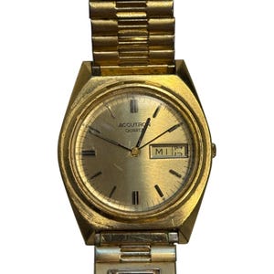 Op de afbeelding: Een goudkleurig Accutron Quartz polshorloge met een rechthoekige wijzerplaat en een goudkleurige band. De wijzerplaat toont de dag en datum. Het horloge heeft een goudkleurige bezel en een goudkleurige band.