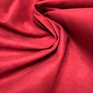 Könnte beinhalten: Nahaufnahme eines tiefroten Stoffes mit einer weichen, Wildleder-ähnlichen Textur. Das Material ist gefaltet und drapiert, wodurch Schatten und Lichtreflexe entstehen, die seine Textur hervorheben. Die Farbe ist ein sattes, gesättigtes Rot.