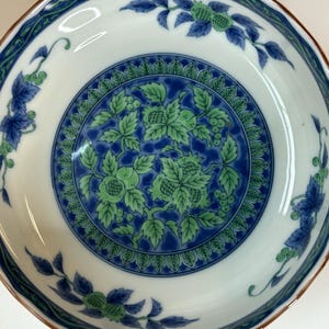 Puede incluir: Un cuenco de cerámica blanco con un diseño floral azul y verde. El cuenco tiene un borde festoneado con un borde marrón. El centro del cuenco presenta un patrón detallado de hojas verdes y detalles azules.