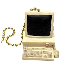 May include: A vintage-style computer ornament with a cream-colored body, black screen, and keyboard. The ornament is attached to a gold-colored beaded chain. The computer has a retro aesthetic, reminiscent of older desktop computers.