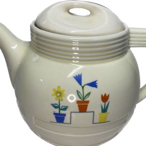 Może przedstawiać: Kremowy ceramiczny imbryk z pokrywką i uchwytem. Imbryk jest ozdobiony kolorowymi kwiatowymi wzorami w doniczkach. Projekt obejmuje żółtą stokrotkę, niebieskie dzwonki i czerwoną tulipan. Imbryk ma dzióbek i okrągłą pokrywkę z otworem.