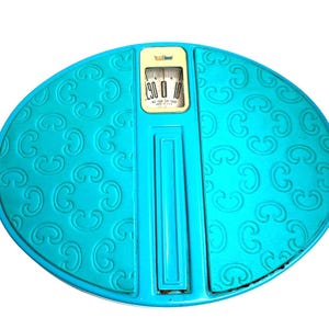 May include: An oval-shaped bathroom scale in a vibrant turquoise color featuring a repeating embossed pattern across the surface. The central display panel is cream-colored with a black analog dial and the text Not Legal For Trade Made In U.S.A.