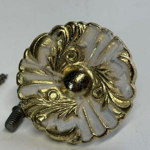 May include: A decorative cabinet knob with a white and gold finish. The knob features an ornate, floral design with a central, dark-colored accent. Two screws are visible, suggesting it's ready for installation.
