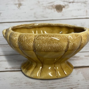 Op de afbeelding: Een vintage kom in sokkelstijl met een geschulpte vormgeving. De kom heeft een warme, gespikkelde geelbruine kleur. Het heeft een afgerond lichaam met een uitlopende basis en twee kleine handvatten. De kom is waarschijnlijk van keramiek of aardewerk.