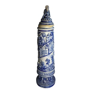 Pode incluir: Um jarro de cerveja alto de cerâmica azul e branca com um design de relevo detalhado. O jarro apresenta uma figura sentada na tampa e cenas de pessoas e texto ao redor do corpo. A base tem um padrão decorativo.