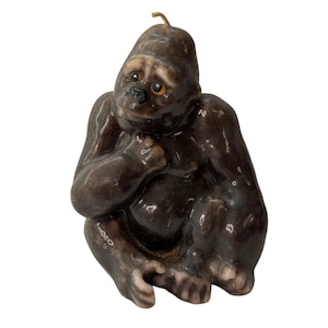 May include: A brown gorilla-shaped candle with a thoughtful expression. The candle has a smooth, glossy finish and a wick at the top. The gorilla is sitting with its legs crossed and its hand on its chin. The candle is a decorative item.