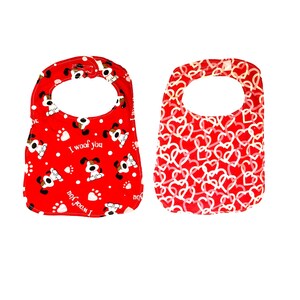 Puede incluir: Dos baberos para bebés. Uno es rojo con huellas de patas blancas, perros de dibujos animados y el texto "I woof you". El otro es rojo con un patrón de corazones blancos. Ambos baberos tienen una parte superior redondeada con un agujero para el cuello.