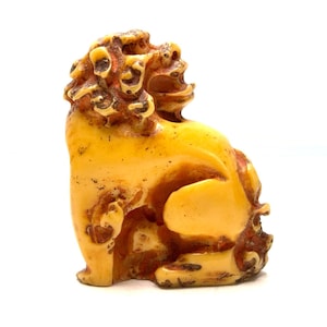 May include: A carved, antique-style figurine of a mythical creature, possibly a lion or dog. The sculpture is primarily a light yellow colour with brown accents, and it is set against a white background. The creature is in a seated position.