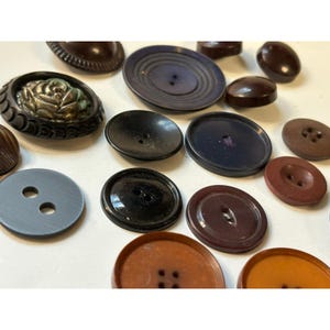 May include: A collection of various vintage buttons in shades of brown, black, and blue. The buttons vary in size, shape, and design, including round, oval, and decorative styles. Some buttons have two or four holes, while others have a shank.