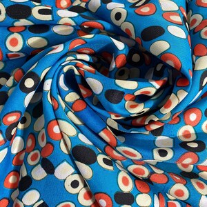 Könnte beinhalten: Ein leuchtend blauer Stoff mit einem sich wiederholenden Muster aus abstrakten Formen in Rot, Schwarz, Weiß und Creme. Der Stoff ist gefaltet, wodurch Schatten und Texturen entstehen. Das Design erinnert an moderne Kunst aus der Mitte des Jahrhunderts.