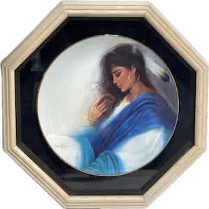 Op de afbeelding: Een decoratieve bord met een portret van een vrouw met lang donker haar, gekleed in een blauw gewaad en gouden oorringen. De plaat heeft een gouden rand en is geplaatst in een achthoekige houten lijst met een zwarte passe-partout.