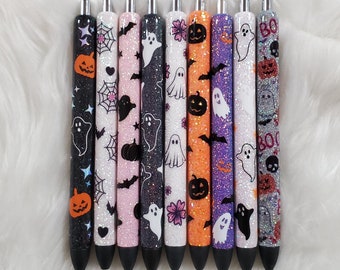 May include: Ten black and silver glitter pens with Halloween themed designs. The designs include ghosts, pumpkins, bats, and spiderwebs. The pens are arranged in a row on a white fluffy surface.