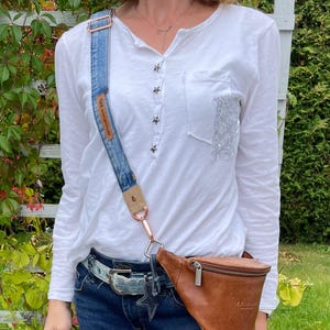 Könnte beinhalten: Eine braune Leder-Bauchtasche mit Reißverschluss und einer silbernen Schnalle. Die Bauchtasche wird über einer blauen Jeans mit einem Jeansgurt getragen. Der Gurt ist mit einem blau-weißen Jeansmuster und einem braunen Lederpatch mit dem Text "Made with love in Amsterdam" verziert.