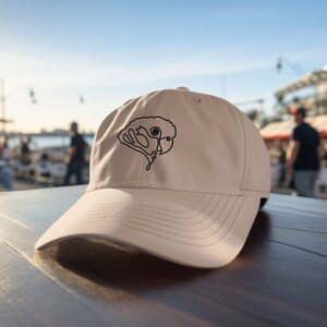 May include: A beige baseball cap with a black line drawing of a face on the front. The cap is sitting on a wooden table outdoors, with a blurred background of people and buildings.