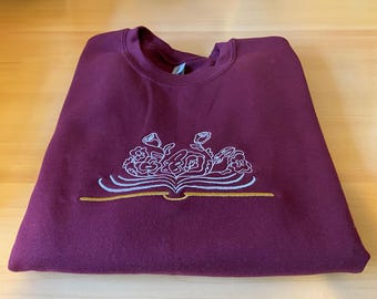 Puede incluir: Sudadera color burdeos con un diseño bordado en blanco de flores y un libro. El diseño está centrado en la parte delantera de la sudadera.