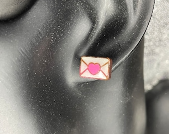 May include: A pair of small wooden earrings shaped like an envelope with a pink heart inside. The earrings are on a mannequin's ear.