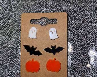 May include: A set of six Halloween-themed earrings featuring two white ghosts, two black bats, and two orange pumpkins. The earrings are displayed on a brown card.