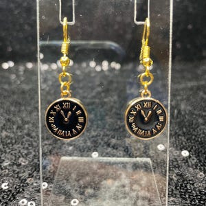 May include: Gold clock face earrings with black Roman numeral markings and gold hands. The earrings are hanging from gold hooks.