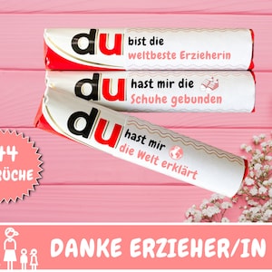 Könnte beinhalten: Drei Schokoladentafeln mit dem Wort "du" darauf gedruckt. Jede Tafel hat eine andere Botschaft darauf gedruckt. Die erste Tafel sagt "du bist die weltbeste Erzieherin". Die zweite Tafel sagt "du hast mir die Schuhe gebunden". Die dritte Tafel sagt "du hast mir die Welt erklärt". Die Tafeln liegen auf einer rosafarbenen Holzoberfläche. Im Hintergrund befindet sich ein weißer Blumenstrauß. Ein rosafarbener Stern-Aufkleber mit dem Text "44 SPRÜCHE" befindet sich in der linken unteren Ecke. Ein rosafarbenes Banner mit dem Text "DANKE ERZIEHER/IN" befindet sich am unteren Rand des Bildes.