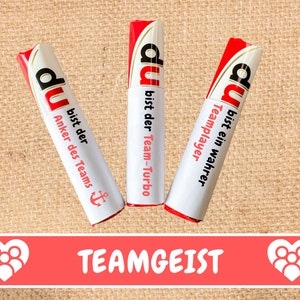 May include: Three red and white wrapped candy bars with the word "du" and different motivational messages in German. The text on the candy bars reads "du bist der Team-Turbo", "du bist ein wahrer Teamplayer", and "du bist der Anker des Teams". The candy bars are on a brown burlap background. The text "TEAMGEIST" is printed in red on a pink background below the candy bars.