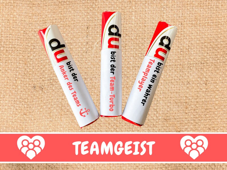 May include: Three red and white wrapped candy bars with the word "du" and different motivational messages in German. The text on the candy bars reads "du bist der Team-Turbo", "du bist ein wahrer Teamplayer", and "du bist der Anker des Teams". The candy bars are on a brown burlap background. The text "TEAMGEIST" is printed in red on a pink background below the candy bars.
