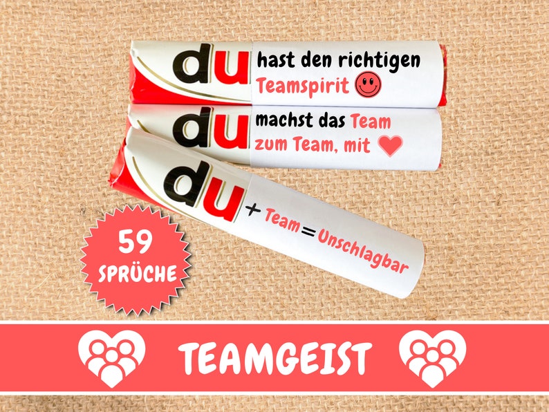 May include: Three white chocolate bars with red and gold accents. The bars have the word "du" printed on them in black. The text on the bars reads "du hast den richtigen Teamspirit", "du machst das Team zum Team, mit", and "du + Team = Unschlagbar". A red starburst sticker with the text "59 SPR&Uuml;CHE" is in the lower left corner. A red banner with the text "TEAMGEIST" and two white heart icons with people inside is at the bottom of the image.