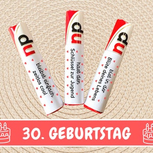 K&ouml;nnte beinhalten: Drei rote und wei&szlig;e Schokoladenriegel mit dem Wort "du" darauf. Jeder Riegel hat eine andere Botschaft. Die Botschaften sind "hast den Schl&uuml;ssel zur Jugend", "bleibst einfach zeitlos cool" und "bist in der Bl&uuml;te deines Lebens". Die Riegel sind in einer Reihe auf einem beigefarbenen Hintergrund angeordnet. Der Text "30. GEBURTSTAG" ist in roten Buchstaben unter den Riegeln geschrieben. Auf beiden Seiten des Textes befinden sich zwei wei&szlig;e Kuchen-Icons mit Kerzen.