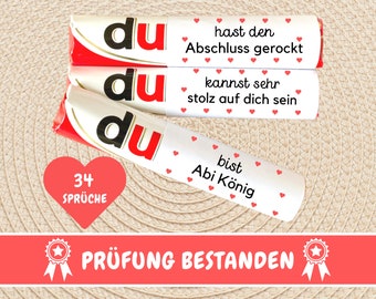 May include: Three white chocolate bars with red hearts and the text "du" on each bar. The bars have different messages on them: "hast den Abschluss gerockt", "kannst sehr stolz auf dich sein", and "bist Abi König". A red heart with the text "34 Sprüche" is below the bars. A red banner with the text "PRÜFUNG BESTANDEN" and two gold stars is below the heart.