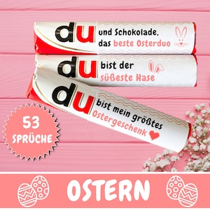 Op de afbeelding: Drie witte chocolade repen met het woord "du" in het zwart op elke reep. De repen hebben verschillende paasberichten erop gedrukt. De repen liggen op een roze houten oppervlak met een witte bloemenarrangement op de achtergrond. Een roze stervormige sticker met de tekst "53 SPRÜCHE" bevindt zich op de voorgrond. De tekst "OSTERN" is in het wit op een roze achtergrond gedrukt met twee witte paaseieren.