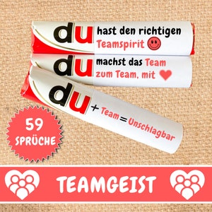 Könnte beinhalten: Drei weiße Schokoladenriegel mit roten und goldenen Akzenten. Die Riegel haben das Wort "du" in Schwarz aufgedruckt. Der Text auf den Riegeln lautet "du hast den richtigen Teamspirit", "du machst das Team zum Team, mit" und "du + Team = Unschlagbar". Ein roter Stern-Aufkleber mit dem Text "59 Sprüche" befindet sich in der linken unteren Ecke. Ein rotes Banner mit dem Text "TEAMGEIST" und zwei weißen Herz-Icons mit Personen darin befindet sich am unteren Rand des Bildes.