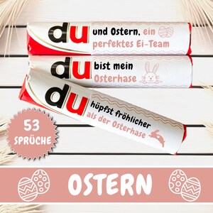 May include: Three white chocolate bars with red and pink accents. The bars have the word "du" printed on them in black. The text on the bars reads "du und Ostern, ein perfektes Ei-Team", "du bist mein Osterhase", and "du hüpfst fröhlicher als der Osterhase". A pink starburst sticker with the text "53 SPRÜCHE" is in the lower left corner. The background is a white wooden surface with a light brown palm frond on the right side.