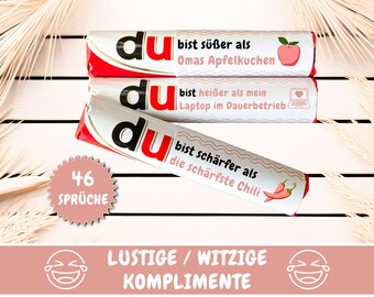 May include: Three red and white candy bar wrappers with the word "du" and a funny compliment on each. The wrappers are stacked on top of each other. The text on the wrappers reads: "du bist süßer als Omas Apfelkuchen", "du bist heißer als mein Laptop im Dauerbetrieb", and "du bist schärfer als die schärfste Chili". There is a pink circle with the text "46 Sprüche" below the wrappers. The text "LUSTIGE/WITZIGE KOMPLIMENTE" is at the bottom of the image.