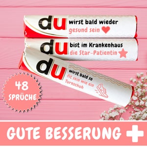 Könnte beinhalten: Drei weiße Schokoladenriegel mit roter und schwarzer Schrift, die "du" und verschiedene Genesungswünsche zeigen. Die Riegel liegen auf einem rosa Hintergrund mit einem weißen Blumenarrangement. Der Text auf den Riegeln lautet "du wirst bald wieder gesund sein", "du bist im Krankenhaus die Star-Patientin" und "du wirst bald so fit sein wie ein Turnschuh". Ein rosa Kreis mit weißer Schrift lautet "48 SPRÜCHE". Der Text "GUTE BESSERUNG +" befindet sich am unteren Rand des Bildes.