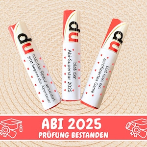 May include: Three white chocolate bars with red and white wrappers. The wrappers have text in German that says "du bist der Abi-Superstar 2025", "du bist nun ein zertifiziertes Genie", and "du hast das Abitur gemeistert jetzt beginnt das Abenteuer". Below the bars is a red banner with white text that says "ABI 2025 PR&Uuml;FUNG BESTANDEN".
