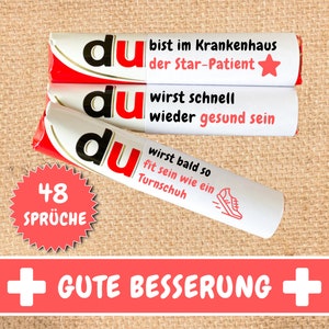 Op de afbeelding: Drie rood-witte verpakte chocolade repen met het woord "du" in zwarte letters en een beterschapsboodschap in het Duits. De repen zijn op elkaar gestapeld. Een rode ster bevindt zich op de bovenste reep. Een rode cirkel met het nummer 48 en het woord "SPRÜCHE" bevindt zich linksonder. De woorden "GUTE BESSERUNG" staan in rode letters op een witte achtergrond met een rood plusteken aan elke kant.