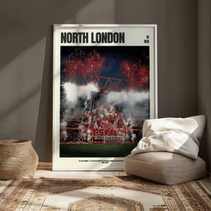 Op de afbeelding: Een ingelijste poster met de tekst "NORTH LONDON IS RED" boven een stadion met rood vuurwerk en een team van spelers. De poster leunt tegen een muur, naast een gevlochten mand en een vloerkussen.