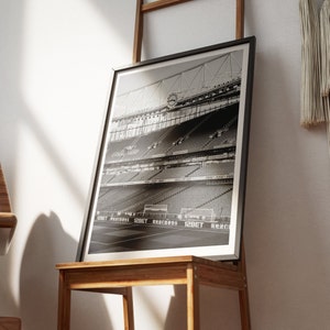Puede incluir: Fotografía en blanco y negro de un gran estadio con filas de asientos. El estadio está vacío y los asientos están todos orientados en la misma dirección. La foto está enmarcada en un marco negro.