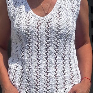May include: White, sleeveless, V-neck top with a delicate, open-weave crochet design. The intricate pattern creates a textured look. The top is made of a soft, lightweight material, perfect for warm weather. A gold necklace is visible.