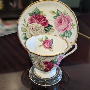 May include: A white teacup and saucer set with a gold rim and floral pattern. The cup and saucer feature pink and white roses with green leaves.