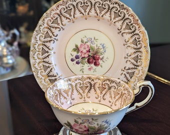 May include: A pink and gold teacup and saucer set with a floral design featuring berries and flowers. The set has a delicate, intricate gold trim.