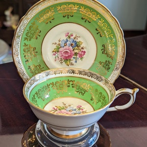 May include: A vintage teacup and saucer set with a green and gold floral design. The cup and saucer have a white background with a green band and gold trim. The floral design features pink, blue, and yellow flowers.