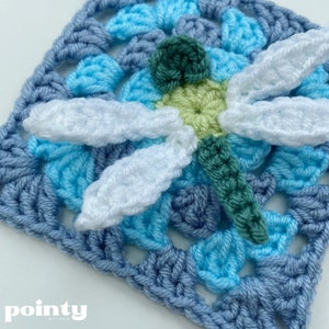 May include: A crocheted square with a blue, light blue, and white color scheme. A white crocheted dragonfly is attached to the center of the square. The dragonfly has green wings and a green body.