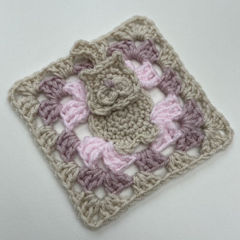 May include: A crocheted square with a pink and beige color scheme. The square features a smaller crocheted flower in the center.