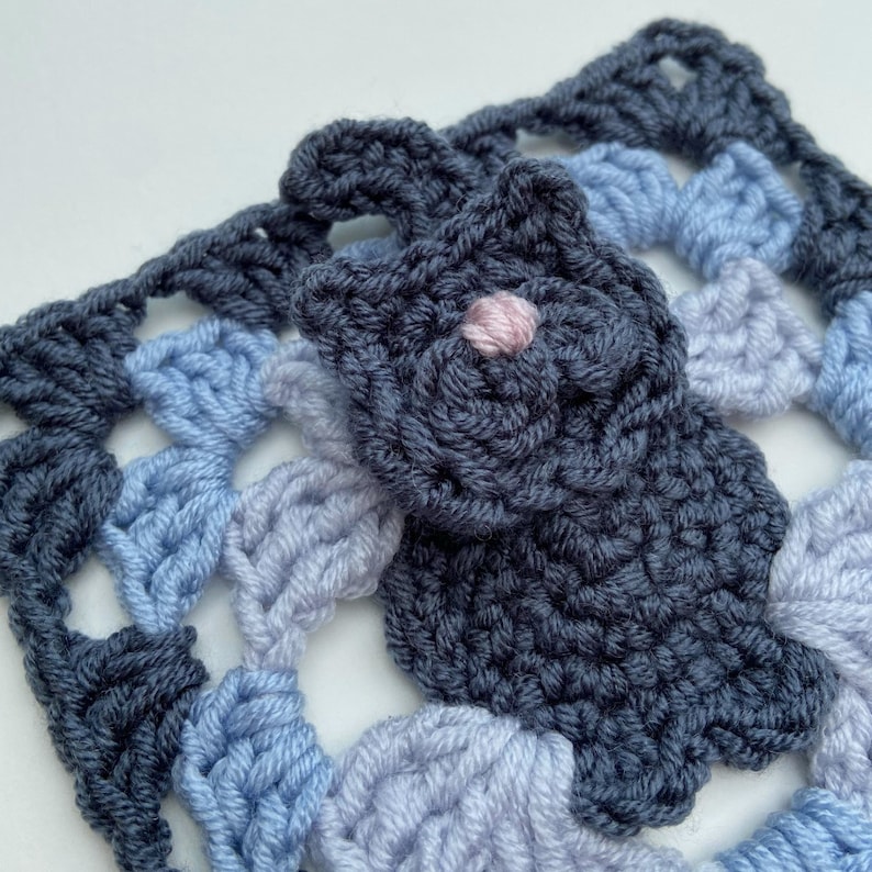 May include: A crocheted granny square with a blue, light blue, and purple color scheme. A dark blue crocheted flower with a pink center is attached to the square.